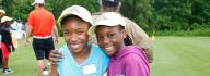 Two girls wearing First Tee — Hampton Roads hats smiling at the camera with golf course in background