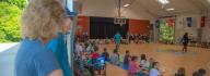 YMCA donors look on at campers sitting on the floor in the gym at YMCA at JT's Camp Grom during a counselor led activity