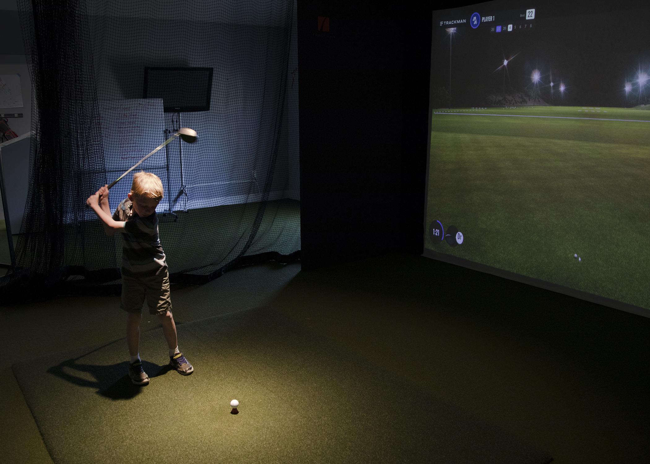 Young boy swings a golf club using TrackMan golf simulator. 