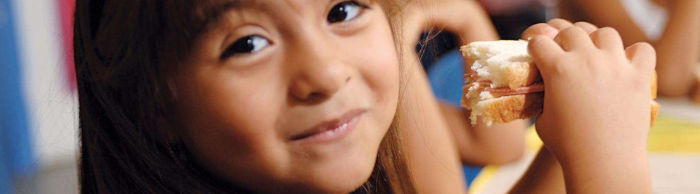 Preschooler smiling at the camera as she eats her sandwich