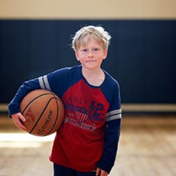 Young caucasian boy holding a YMCA basketball wearing a red shirt with navy long sleeves in an indoor gym basketball court