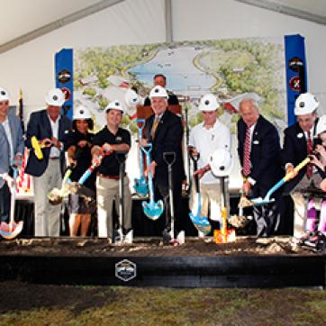Group of people in white hard hats at a ground breaking event with a map of camp grom behind them.