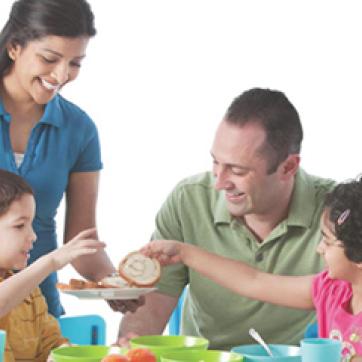 Multi ethnic family of 4 sharing breakfast at a table on a white background.