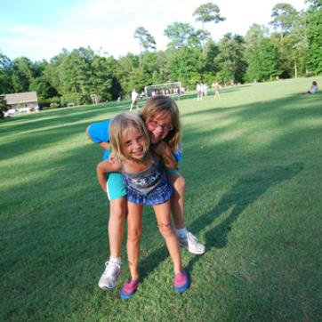 Girl trying to give her sister or friend a piggy back ride in an open soccer field of green grass.