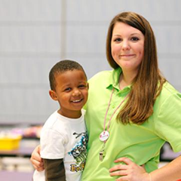African American young child with YMCA female staff in a bright green shirt indoors with students in the background.
