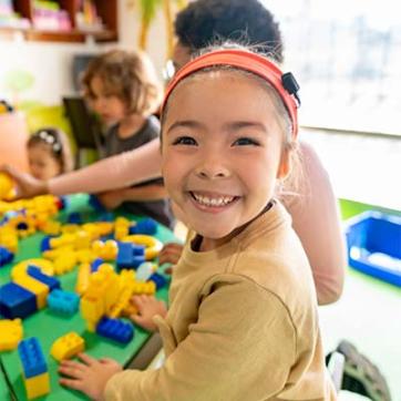 Young girl smiles at the camera in front of a table with blocks, a staff member and other children