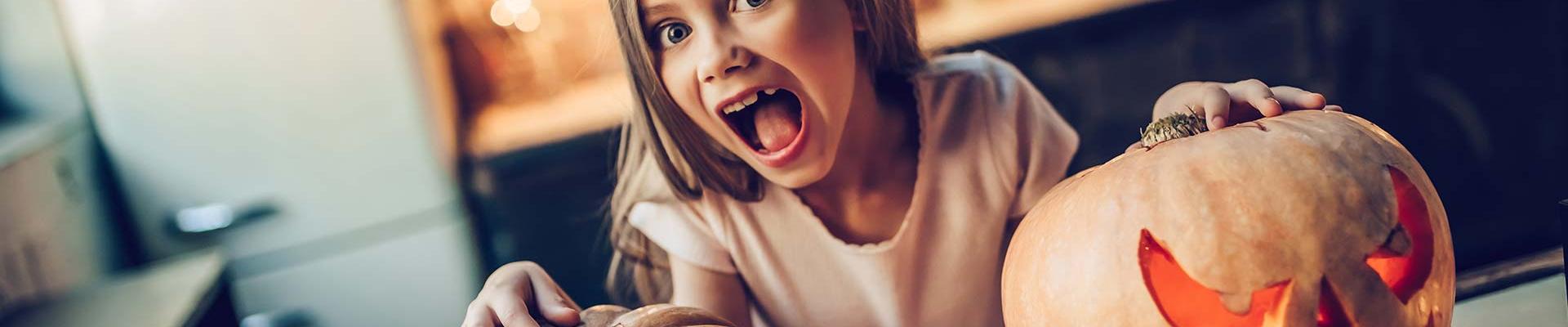 Young, blonde girl standing in front of two pumpkins she carved, pretending to be scared.
