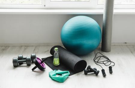 Group of different exercising equipment on white home gym floor. Fitness ball, round foam roller, resistance exercise latex band, jumping rope, dumbbells, yoga mat. 