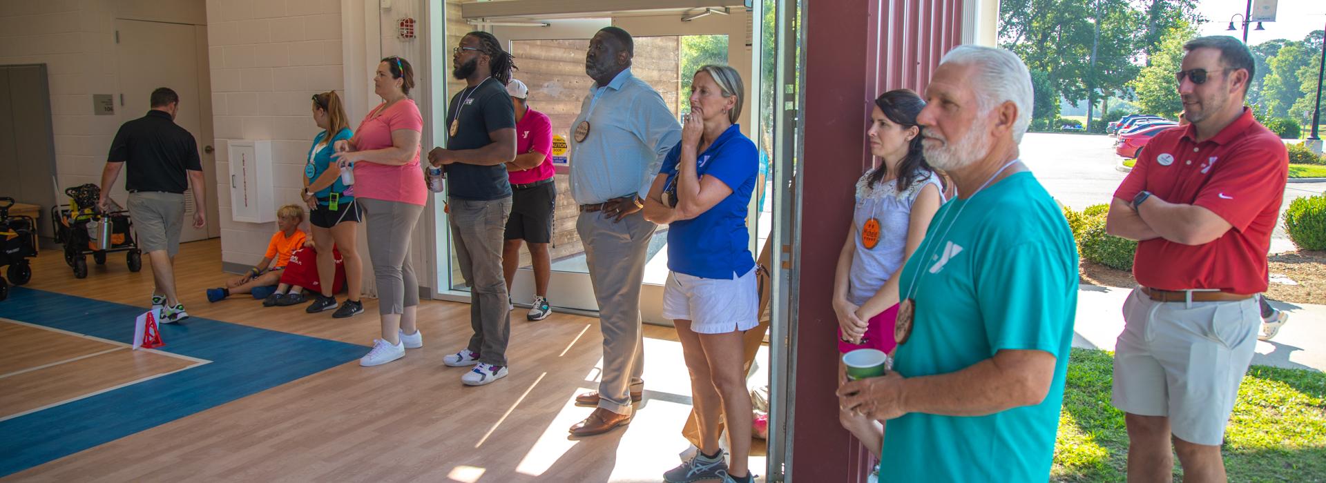 YMCA donors look on at campers sitting on the floor in the gym at YMCA at JT's Camp Grom during a counselor led activity