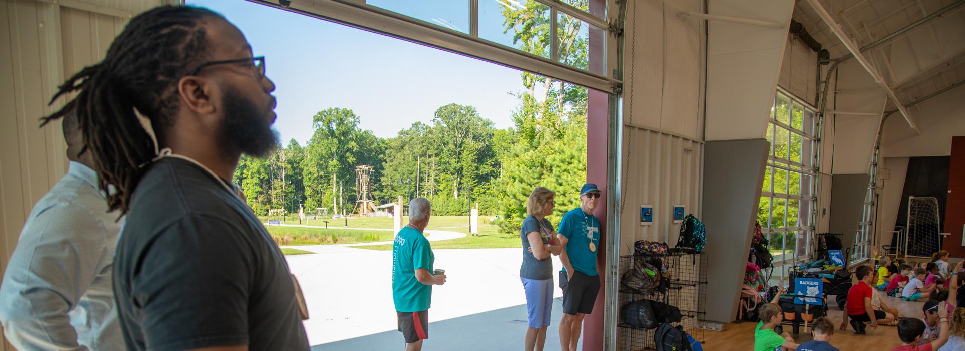 YMCA donors look on at campers sitting on the floor in the gym at YMCA at JT's Camp Grom during a counselor led activity