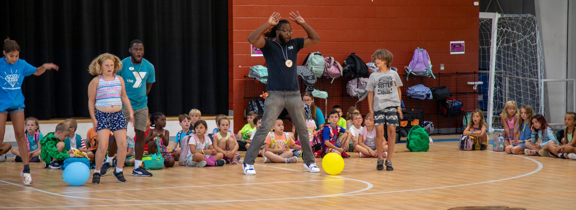 YMCA at JT's Camp Grom counselor leads campers through game in the gymnasium, while other campers sit on the floor watching