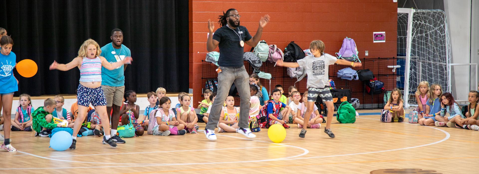 YMCA at JT's Camp Grom counselor leads campers through game in the gymnasium, while other campers sit on the floor watching