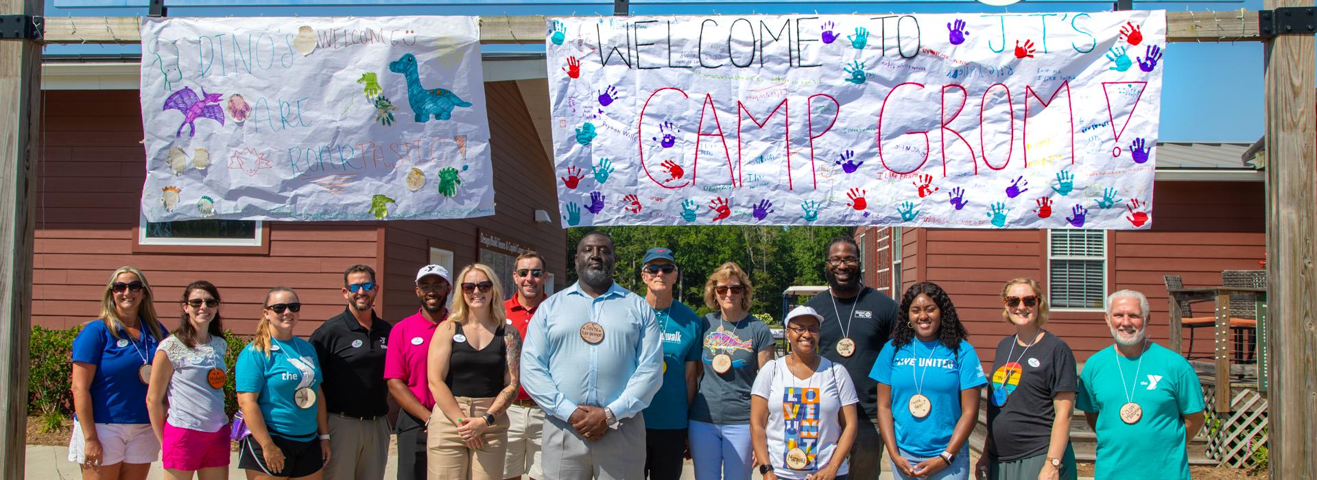 YMCA donors and staff pose under the Welcome to JT's Camp Grom sign at camp entrance