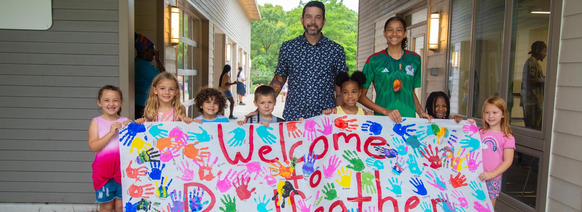 Campers and adults stand in a group holding the Welcome to Camp Red Feather sign