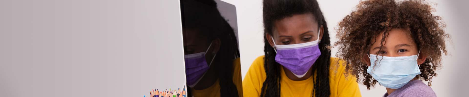 A child in a mask sits with a staff member also in a mask, in front of a computer