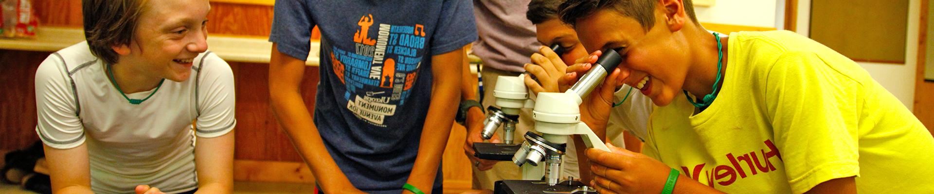 Two campers looking in microscopes while other campers look in, smiling and having fun discovering in science lab at YMCA Camp Silver Beach