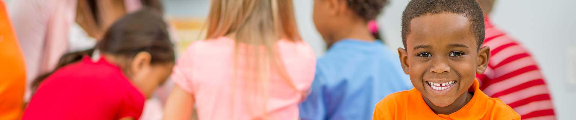 A young boy in an after-school program smiles at us in front of a group of children involved in an activity