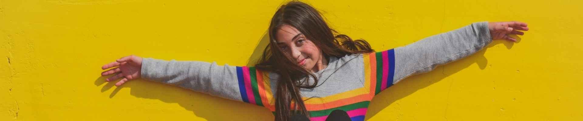 Teen wearing a gray sweater with rainbow stripes sits against yellow wall and smiles while holding arms wide