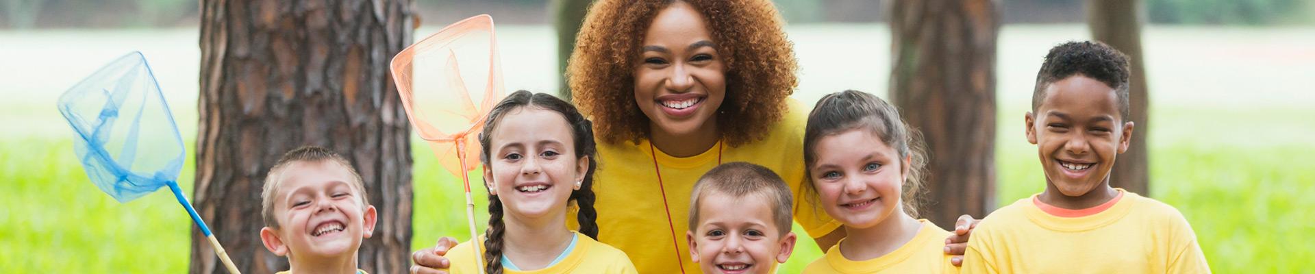 Camp counselor and campers, standing in front of trees and holding butterfly nets, smile at the camera