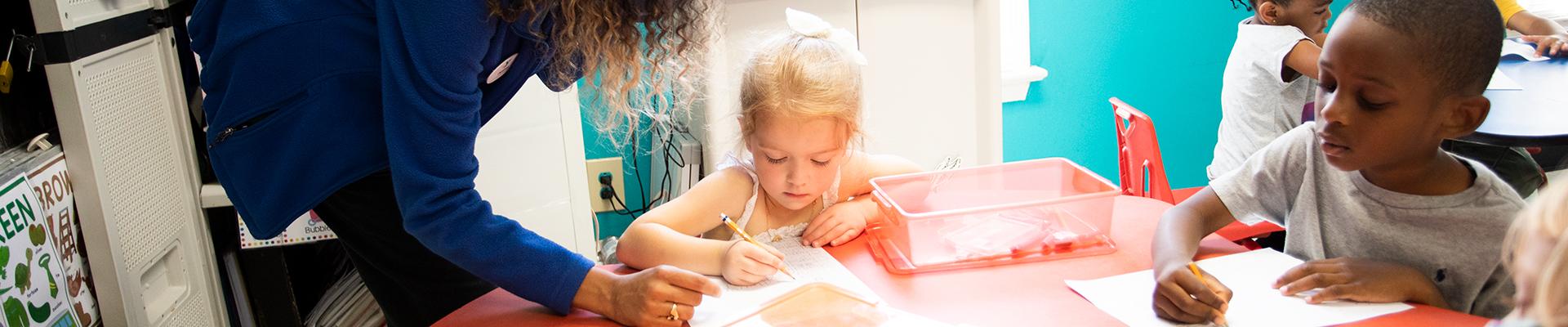 Two young children sit at a table writing, while a teacher assists in YMCA preschool