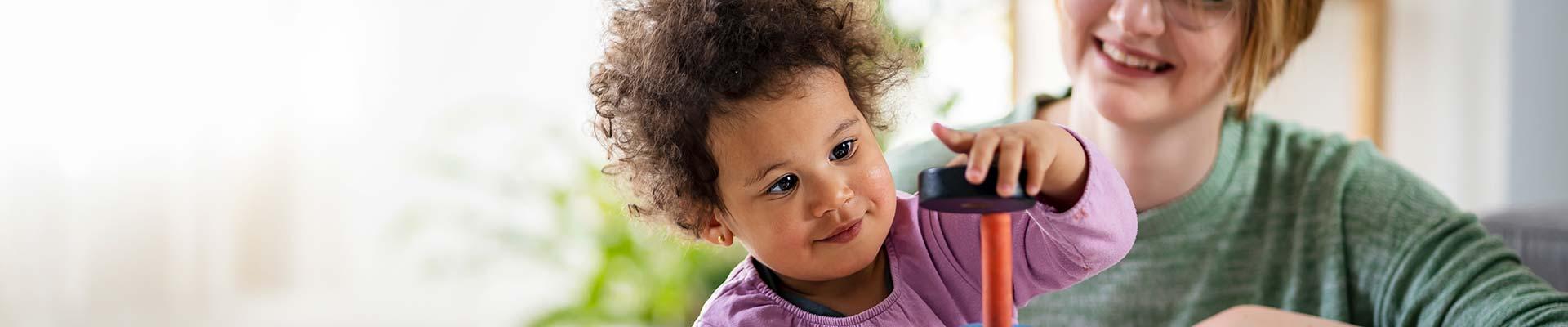 Child playing with a stacking tower while a caregiver looks on, smiling.