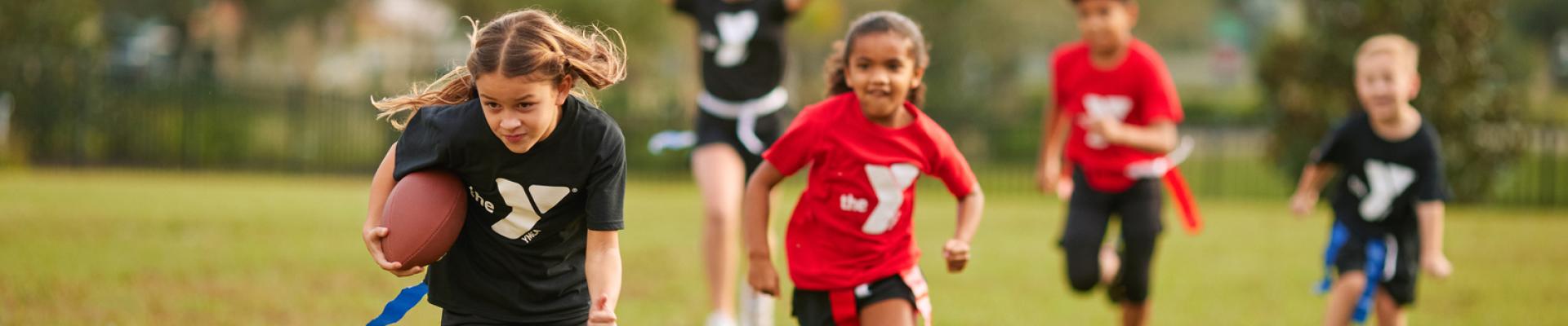 Five teenagers run down the flag football field during an outdoor game with one at the front carrying the football.