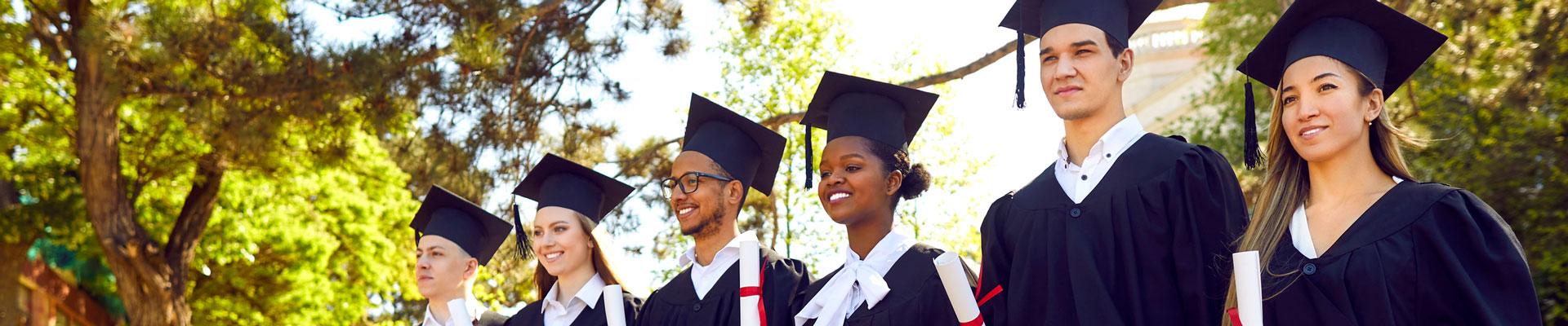 Students in graduation cap and gowns with diplomas standing in a line outside