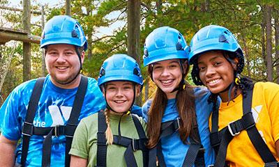 Teen leaders and staff advisor in helmets and safety gear prepare to zipline in adventure park