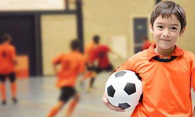 Young child smiles at the camera while holding a soccer ball. In the background other kids run around playing.