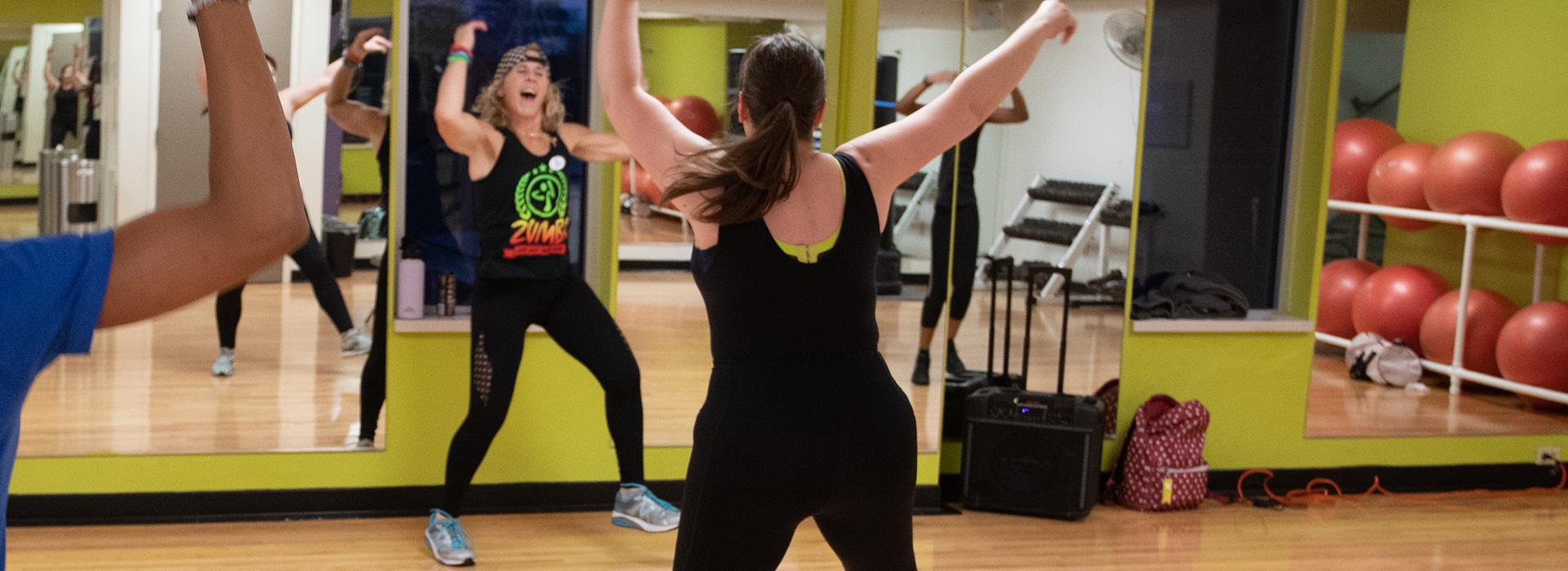 An instructor teaches an exciting Zumba class with arms up in the air and joy on her face.