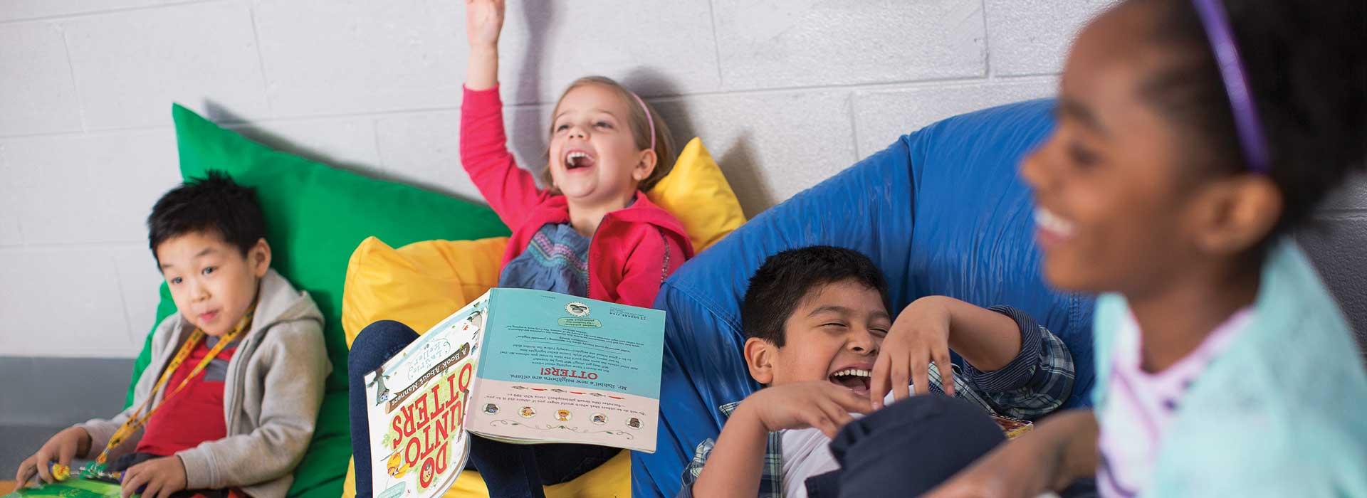 Kids in YMCA child care, resting against bean bag pillows with books, laughing and having fun