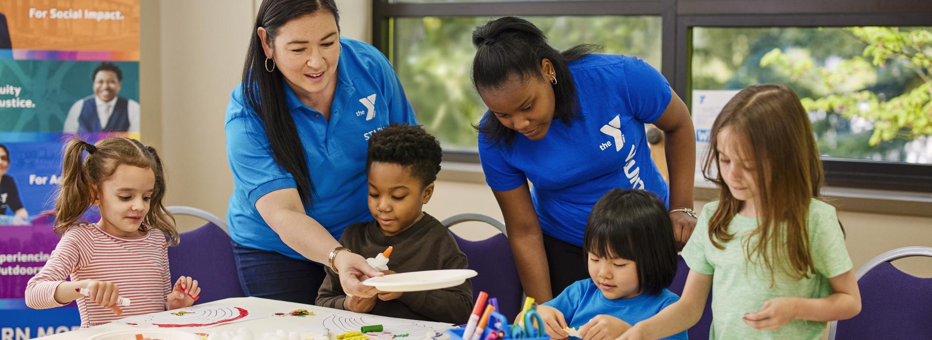 Two YMCA staff members help four children with arts and crafts project in YMCA childcare.