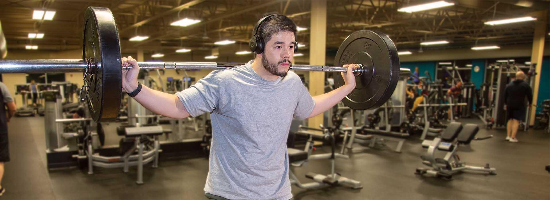 Male holding a barbell with plates on his shoulders, headphones on, working out at the Y